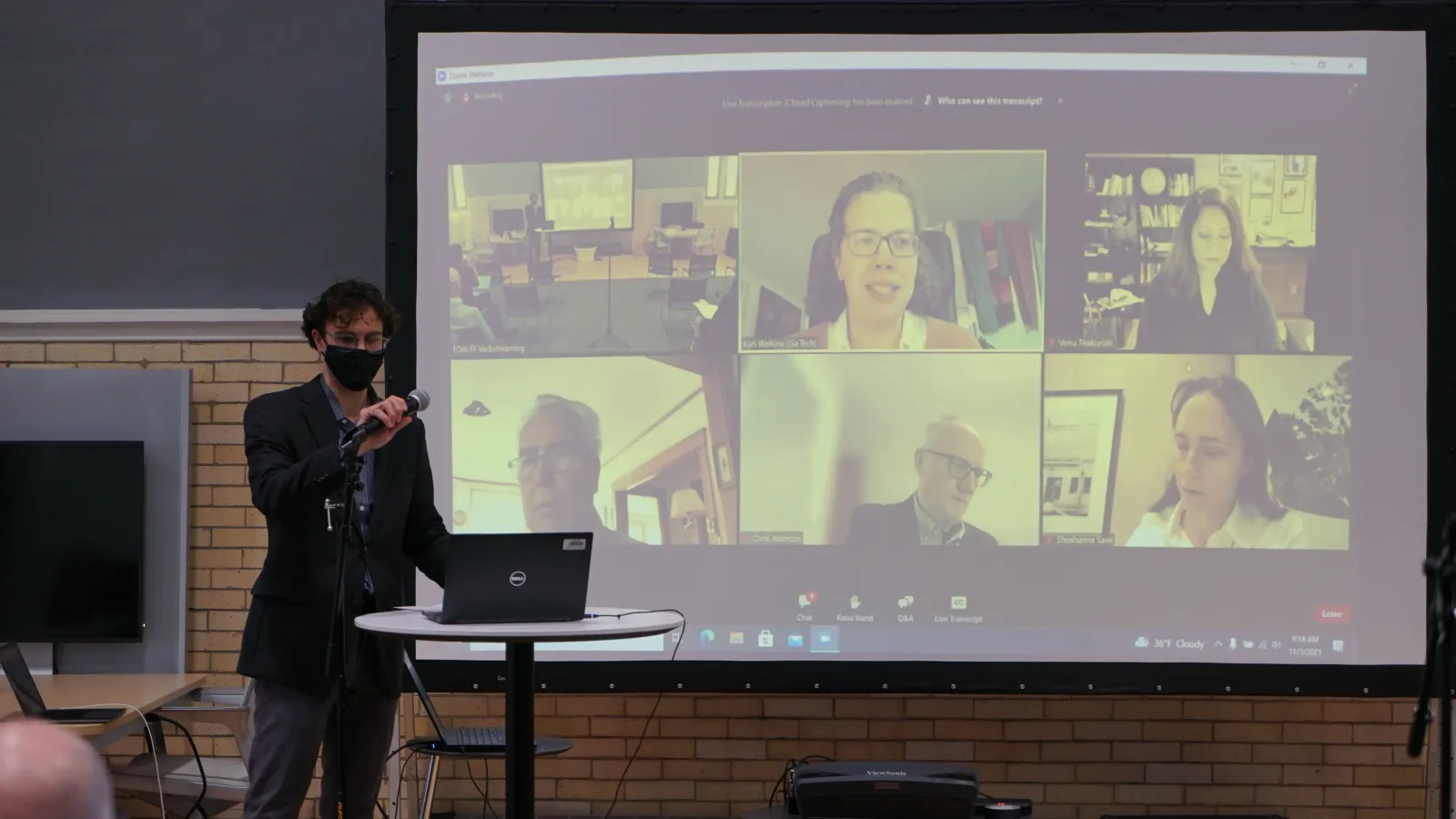 A man moderates a discussion in front of an audience as panelists on Zoom display on a screen behind him