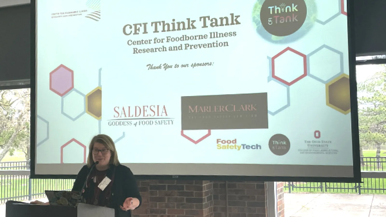 Dr. Barb Kowalcyk speaking at a podium in front of a slide show presentation