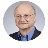 Headshot of Dr. Ray Perrault, wearing glasses, a dark blazer, and a blue shirt, against a gray background.