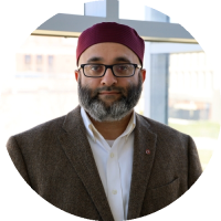 Headshot of Ayaz Hyder wearing glasses and a jacket, standing indoors.