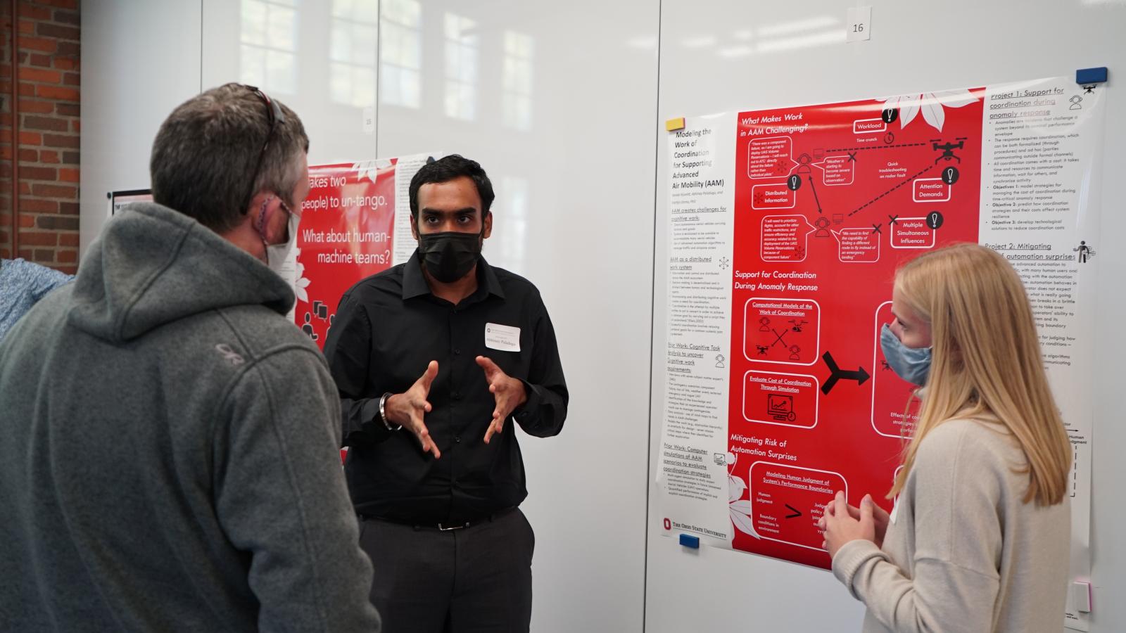 A man discusses his research poster with another man and a woman at the Fall Forum poster session