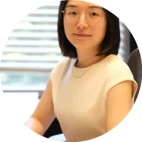 Headshot of Xian Yu wearing glasses and a light top, photographed indoors with horizontal window blinds in the background.